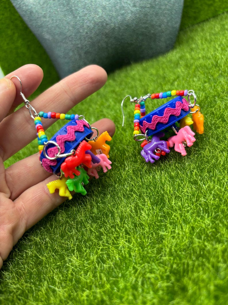 May include: A pair of dangle earrings featuring a blue plastic bottle cap base decorated with pink rickrack trim. The earrings are suspended by multi-colored rainbow beads and feature small, colorful plastic horse charms in shades of red, yellow, green, orange, and purple hanging from the bottom.