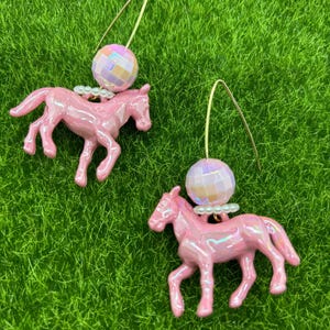 May include: A pair of dangle earrings featuring a glossy pink horse figurine suspended from a gold-toned wire hook. Above each horse is a small white bead spacer and a round, iridescent, faceted disco ball bead that reflects light.