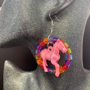 May include: A dangle earring featuring a glossy pink horse charm suspended within a circular hoop made of translucent, multi-colored plastic pony beads. The earring is attached to a silver-toned metal fishhook style ear wire for pierced ears.