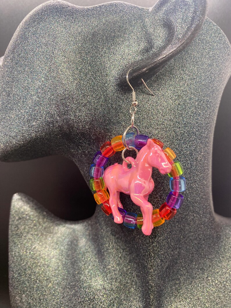 May include: A dangle earring featuring a glossy pink horse charm suspended within a circular hoop made of translucent, multi-colored plastic pony beads. The earring is attached to a silver-toned metal fishhook style ear wire for pierced ears.