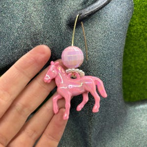 May include: A dangle earring featuring a glossy, iridescent pink horse charm suspended from a small ring of white beads. Above the horse is a faceted, light pink spherical bead attached to a thin, gold-toned metal ear wire hook.