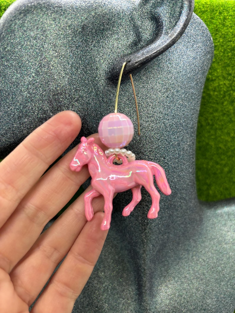May include: A dangle earring featuring a glossy, iridescent pink horse charm suspended from a small ring of white beads. Above the horse is a faceted, light pink spherical bead attached to a thin, gold-toned metal ear wire hook.
