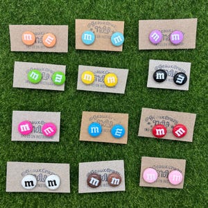以下が含まれることがあります： 「M」と「E」の文字が書かれた、カラフルな丸いキャンディーモチーフのイヤリングの詰め合わせです。イヤリングは、「@GeauxCrazyNails」と「Tag us on Instagram!」の文字が書かれた小さなカードに飾られています。