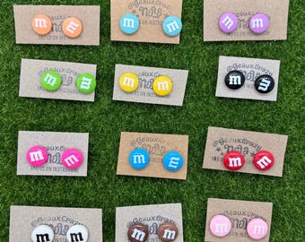 May include: Assortment of colorful, round, candy-themed earrings with the letters "M" and "E" on them. The earrings are displayed on small cards with the text "@GeauxCrazyNails" and "Tag us on Instagram!"