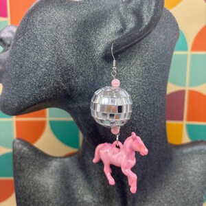 May include: A dangling earring featuring a silver-toned metal hook, a small pink bead, a spherical disco ball made of tiny mirrored glass squares, another pink bead, and a pink plastic horse figurine suspended at the bottom of the earring.