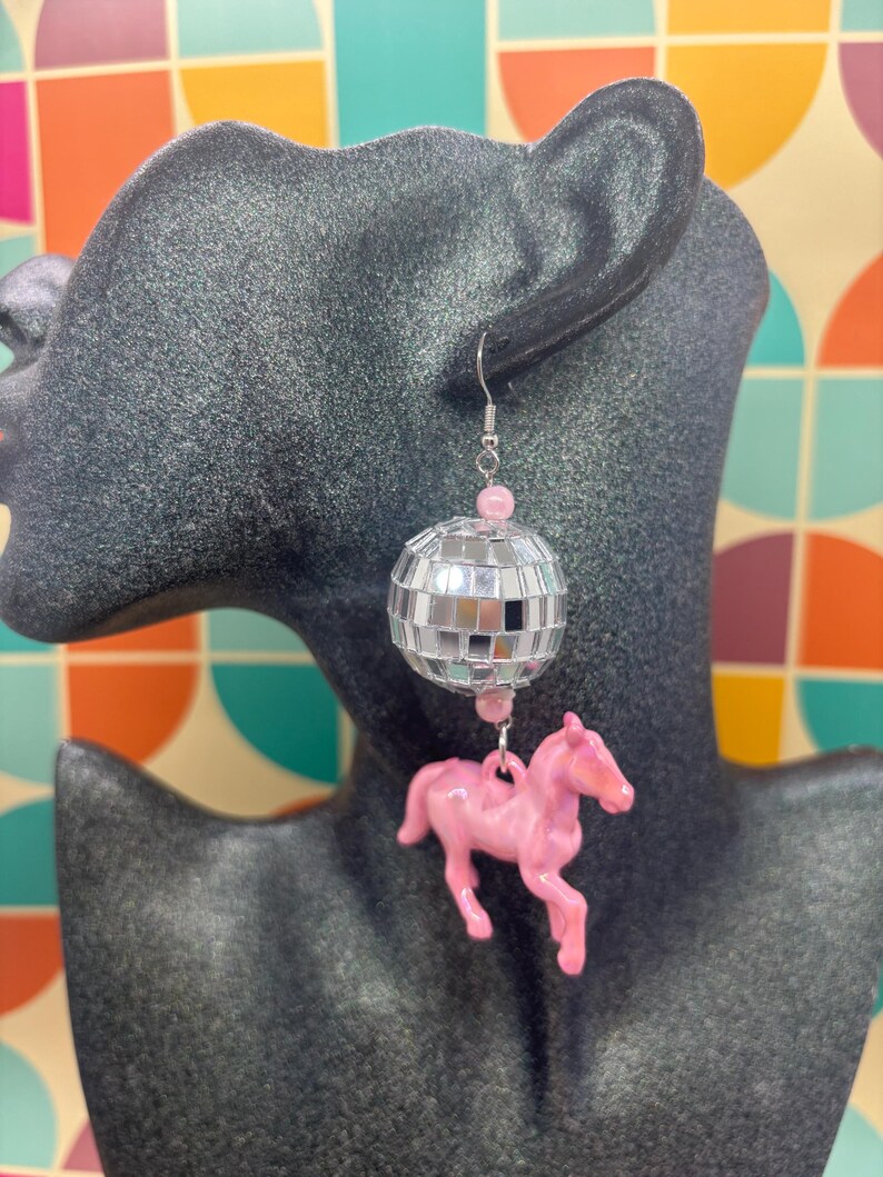May include: A dangling earring featuring a silver-toned metal hook, a small pink bead, a spherical disco ball made of tiny mirrored glass squares, another pink bead, and a pink plastic horse figurine suspended at the bottom of the earring.