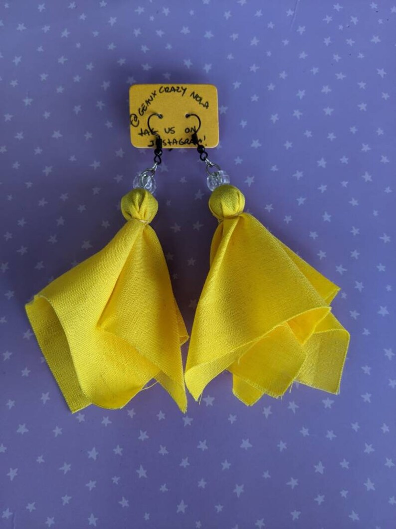 Puede incluir: Un par de pendientes de tela amarilla con cuentas transparentes y ganchos negros. Los pendientes cuelgan de una tarjeta que dice "@genix_crazy_nola follow us on instagram!"