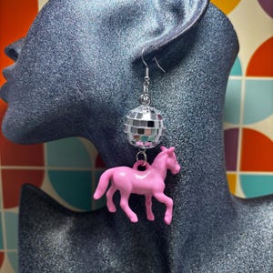 May include: A dangling earring featuring a small, reflective silver disco ball charm suspended above a solid pink plastic horse figurine. The earring is attached to a silver-toned metal hook, creating a layered design with a shiny finish.