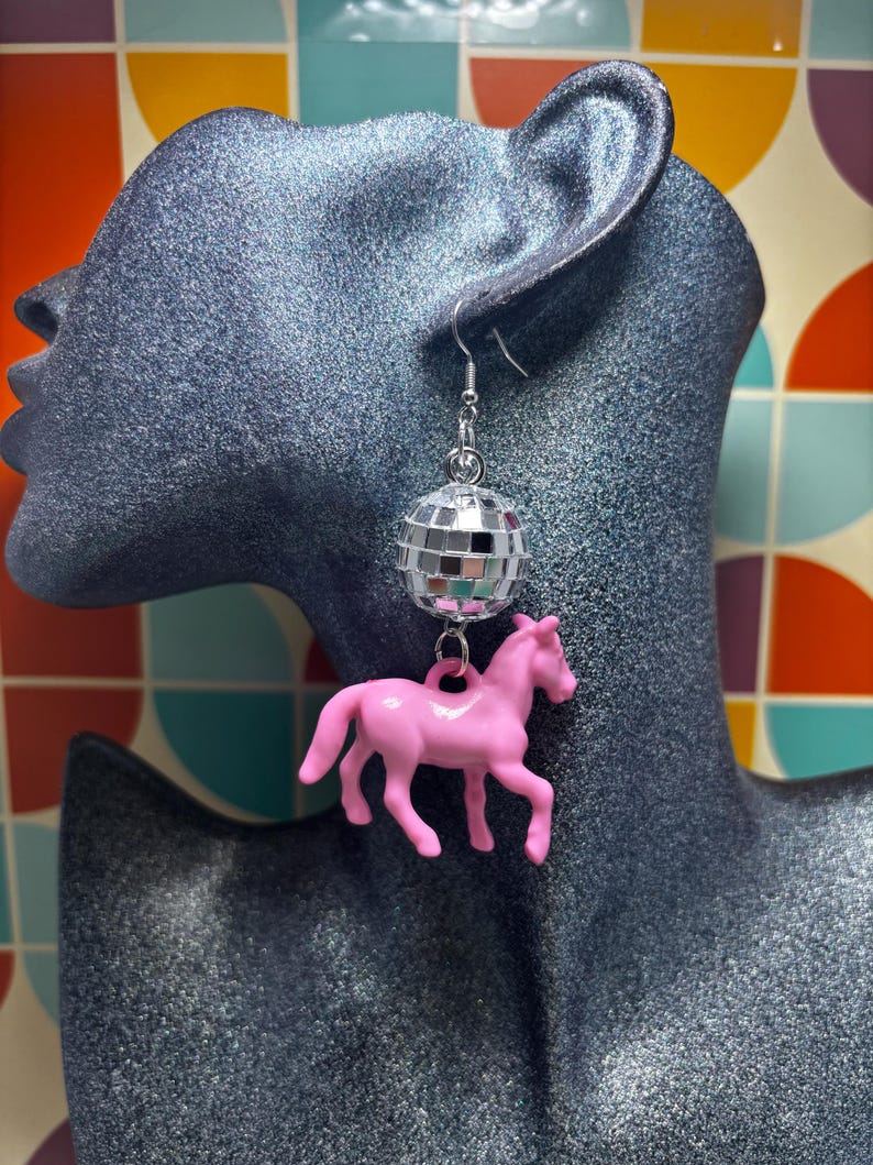 May include: A dangling earring featuring a small, reflective silver disco ball charm suspended above a solid pink plastic horse figurine. The earring is attached to a silver-toned metal hook, creating a layered design with a shiny finish.