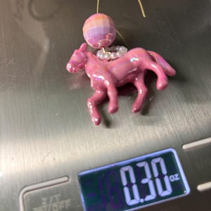 May include: A pink horse-shaped earring charm with an iridescent finish, featuring a small white beaded ring around its neck and a faceted, multi-colored spherical bead above it, all suspended from a gold-tone wire hook, weighing 0.30 oz.
