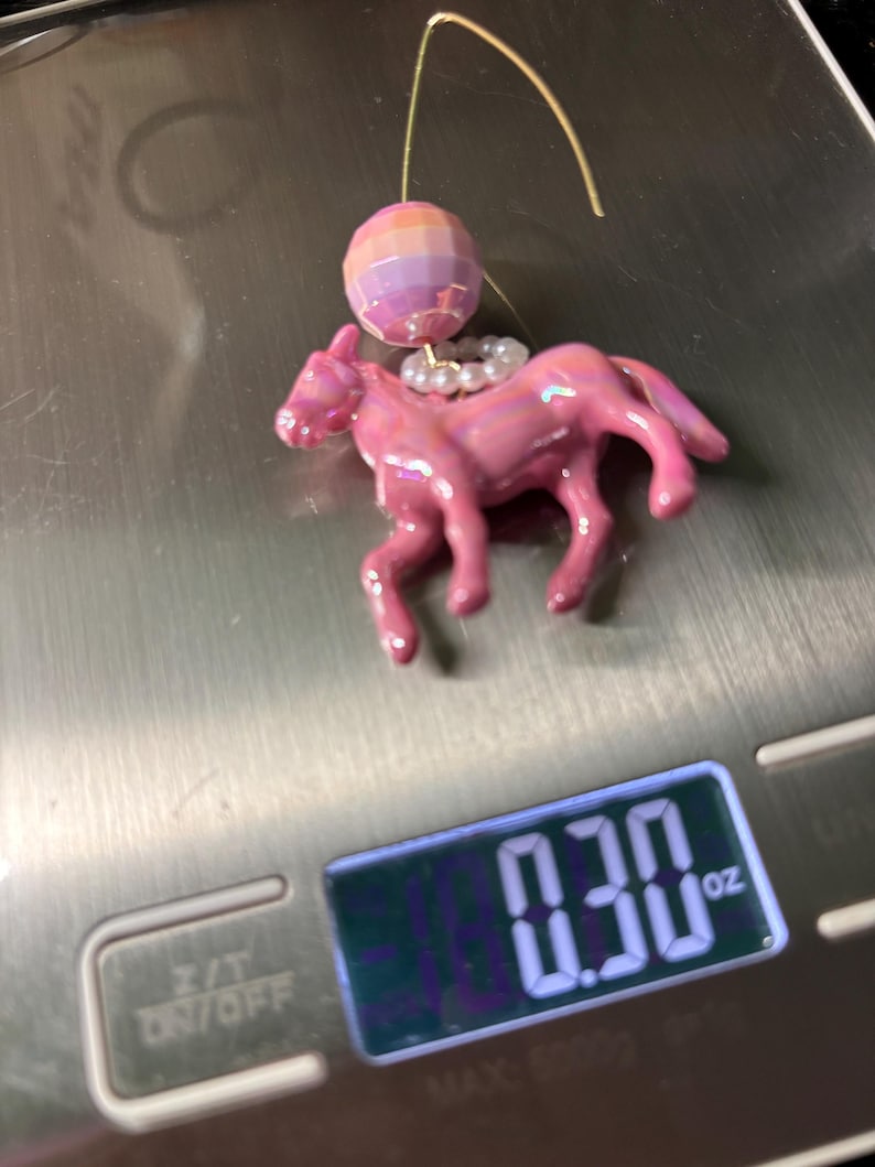 May include: A pink horse-shaped earring charm with an iridescent finish, featuring a small white beaded ring around its neck and a faceted, multi-colored spherical bead above it, all suspended from a gold-tone wire hook, weighing 0.30 oz.