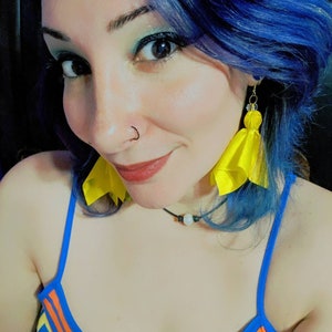Puede incluir: Una mujer con el pelo azul lleva un top de tirantes azul y naranja a rayas y pendientes de borla amarillos.