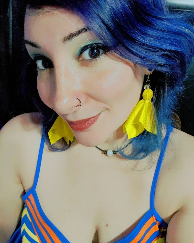 Puede incluir: Una mujer con el pelo azul lleva un top de tirantes azul y naranja a rayas y pendientes de borla amarillos.