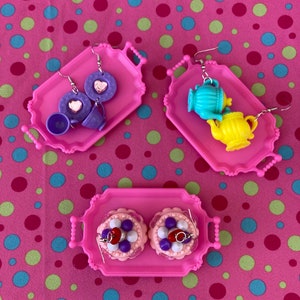 May include: Three pink trays with silver earrings. The first tray has a purple teacup, a purple saucer, and a heart-shaped charm. The second tray has a turquoise teapot and a yellow teapot. The third tray has two pink cakes with purple and red decorations.