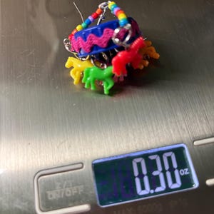 May include: A novelty earring featuring a blue plastic bottle cap base decorated with a pink rickrack trim and a rainbow-colored beaded hoop. Small, colorful plastic horse charms in yellow, green, and red hang from the cap, weighing 0.30 oz.