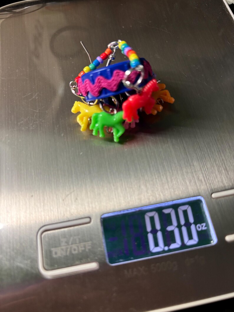 May include: A novelty earring featuring a blue plastic bottle cap base decorated with a pink rickrack trim and a rainbow-colored beaded hoop. Small, colorful plastic horse charms in yellow, green, and red hang from the cap, weighing 0.30 oz.