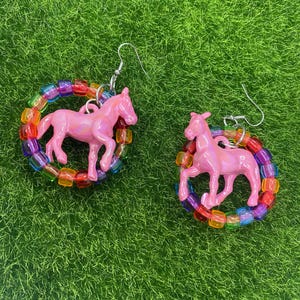 May include: A pair of dangle earrings featuring a glossy pink plastic horse figurine suspended within a circular frame made of translucent, multi-colored square pony beads. The earrings are attached to silver-toned metal fishhook ear wires.