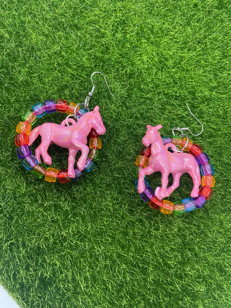 May include: A pair of dangle earrings featuring a glossy pink plastic horse figurine suspended within a circular frame made of translucent, multi-colored square pony beads. The earrings are attached to silver-toned metal fishhook ear wires.