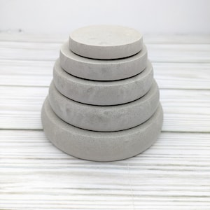 May include: A stack of five gray concrete circular coasters, each with a slightly different diameter, arranged in a pyramid shape on a white wooden surface.