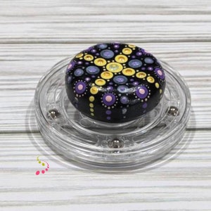 May include: A black stone painted with a mandala design in shades of purple, yellow, and white. The stone is on a clear rotating display stand.
