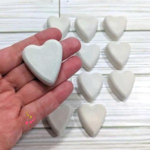May include: A collection of 12 white heart-shaped ceramic ornaments. The hearts are arranged in a grid pattern on a white wooden surface. One heart is being held in a hand.