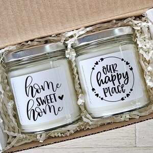 May include: Two white candles in glass jars with silver lids. The candles are in a white box with brown paper shredding. The labels on the candles say "home sweet home" and "our happy place".