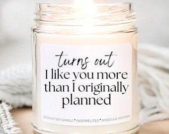 May include: A white candle in a glass jar with a label that reads "turns out I like you more than I originally planned". The candle is lit and the flame is visible.