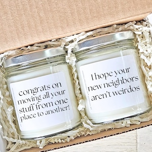 May include: Two glass jar candles with silver lids inside a cardboard box. The candles have white labels with black text that reads, "congrats on moving all your stuff from one place to another!" and "I hope your new neighbors aren't weirdos."