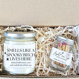 May include: A cardboard gift box containing a glass candle jar and a small glass jar of matches nestled in paper filler. The candle label reads: SMELLS LIKE A SPOOKY BITCH LIVES HERE, infused with sweet, sass and a whole lot of bad ass. The match jar reads: light em up.