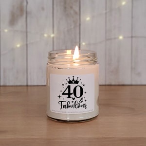 May include: A white candle in a glass jar with a black and white label that says "40 & Fabulous" with a crown and stars. The candle is lit and the flame is visible.
