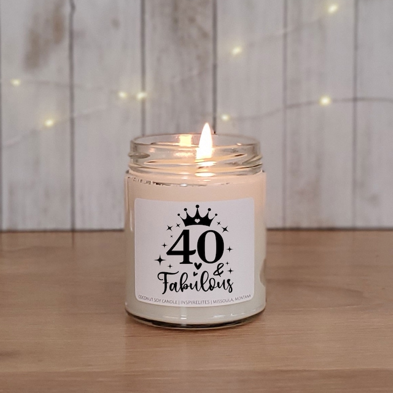 May include: A white candle in a glass jar with a black and white label that says "40 & Fabulous" with a crown and stars. The candle is lit and the flame is visible.