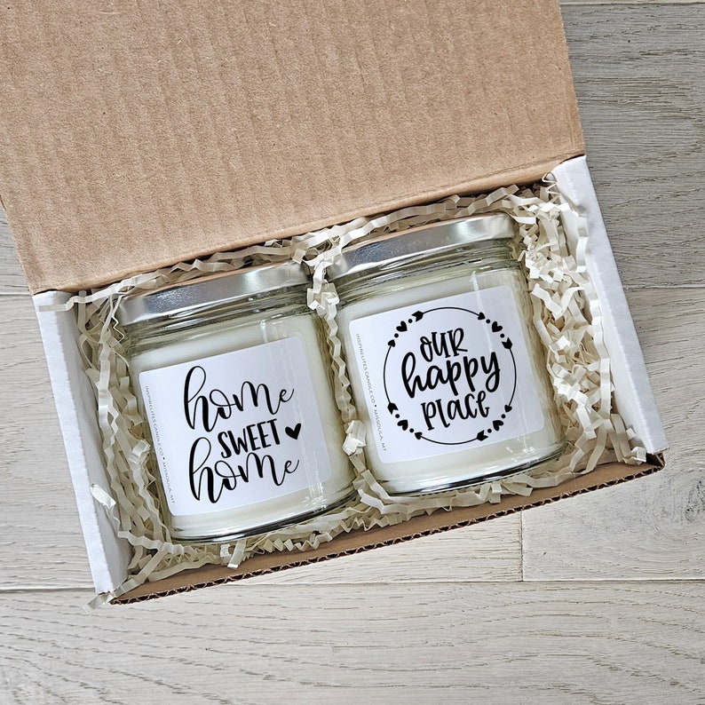 May include: Two white candles in glass jars with silver lids. The candles are in a white box with brown paper shredding. The labels on the candles say "home sweet home" and "our happy place".