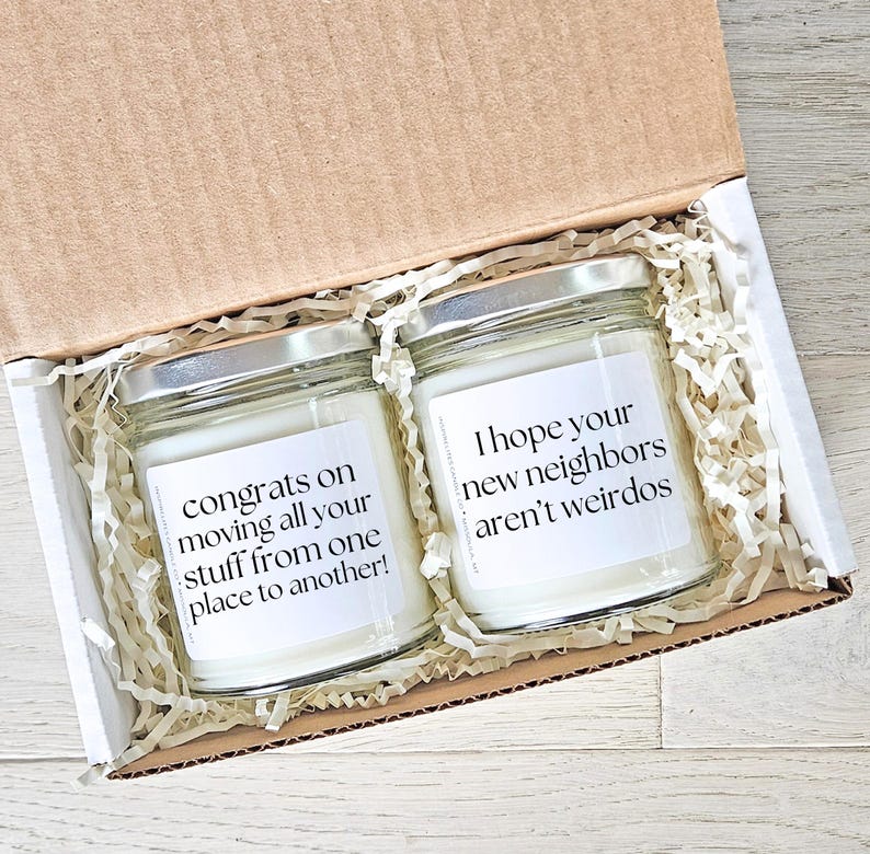 May include: Two glass jar candles with silver lids inside a cardboard box. The candles have white labels with black text that reads, "congrats on moving all your stuff from one place to another!" and "I hope your new neighbors aren't weirdos."