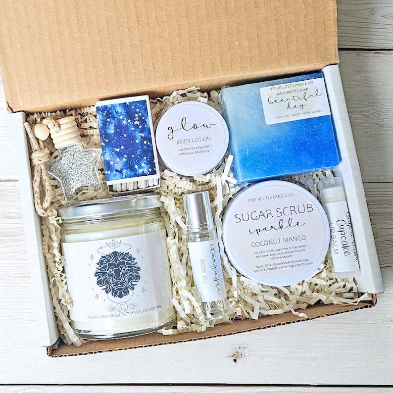 May include: A gift box containing various self-care products. The box includes a candle, body lotion, soap, sugar scrub, lip balm, and body oil. Also present are a star-shaped car air freshener and a box of matches with a starry design.