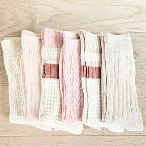 May include: Six pairs of socks in shades of pink, white, and beige, arranged on a light wooden surface. Some socks have ribbed textures, while others feature a striped design with a brown accent.