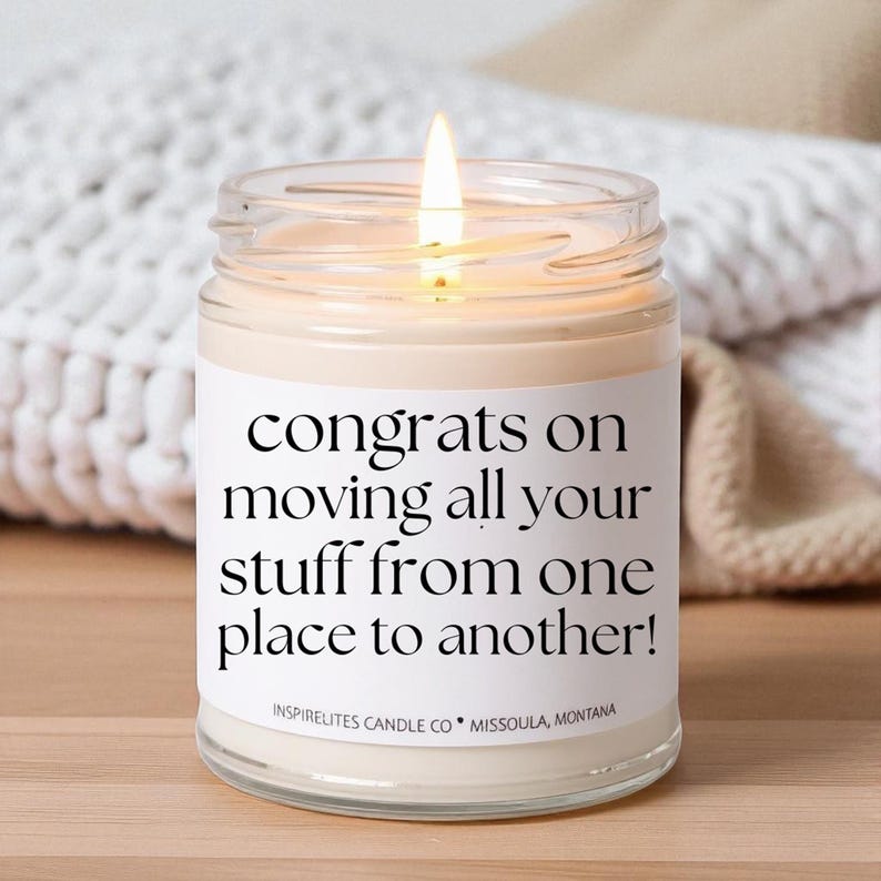 May include: A lit candle in a clear glass jar with a white label. The label reads "congrats on moving all your stuff from one place to another!" The candle is from Inspirelites Candle Co., Missoula, Montana.