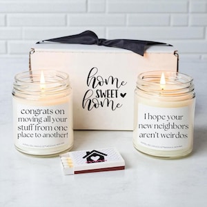 May include: A gift set with two lit jar candles and a matchbox. The candles have labels with humorous messages. A white box with a black bow and the words "home sweet home" is in the background. The matchbox has a house graphic.