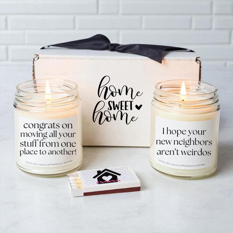 May include: A gift set with two lit jar candles and a matchbox. The candles have labels with humorous messages. A white box with a black bow and the words "home sweet home" is in the background. The matchbox has a house graphic.