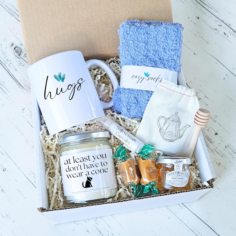 Pu&ograve; includere: Una confezione regalo piena di una tazza bianca con testo "hugs", calzini blu, una candela con testo "at least you don't have to wear a cone", miele, caramelle, balsamo per le labbra, una bustina di t&egrave; e un misurino di miele in legno. La scatola &egrave; foderata di carta.