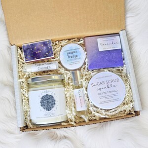 May include: A gift box containing various self-care items. It features a candle, body lotion, sugar scrub, body oil, soap, lip balm, and matches. The products are labelled with the text "Juniper Breeze", "Cashmere", and "Coconut Mango".
