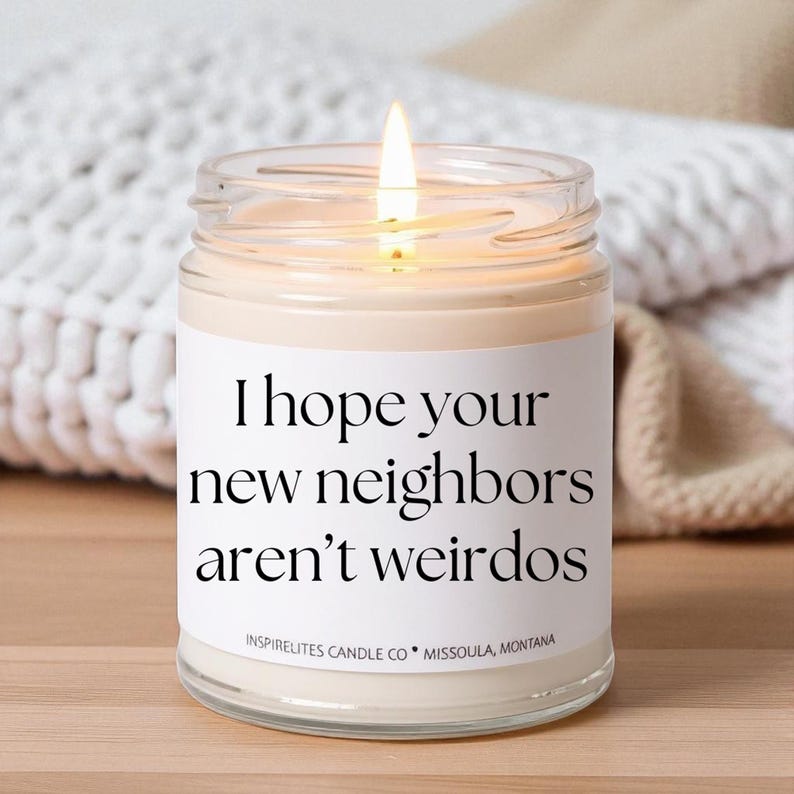 May include: A lit candle in a clear glass jar with a white label. The label reads "I hope your new neighbors aren't weirdos." The candle is from Inspirelites Candle Co., Missoula, Montana. The background includes a white knitted blanket and a beige fabric.