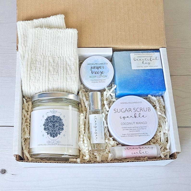 May include: A gift box containing self-care products. It features a candle with a lion design, body lotion, a blue soap bar, sugar scrub, lip balm, a roller bottle, and cream socks. The box is filled with shredded paper.