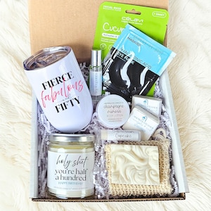 May include: A gift box filled with self-care items. Includes a white tumbler with "Fierce Fabulous Fifty" text, a candle, body lotion, lip balm, eye mask, facial mask, and shower steamers. The candle reads "holy shit you're half a hundred Happy Birthday!"