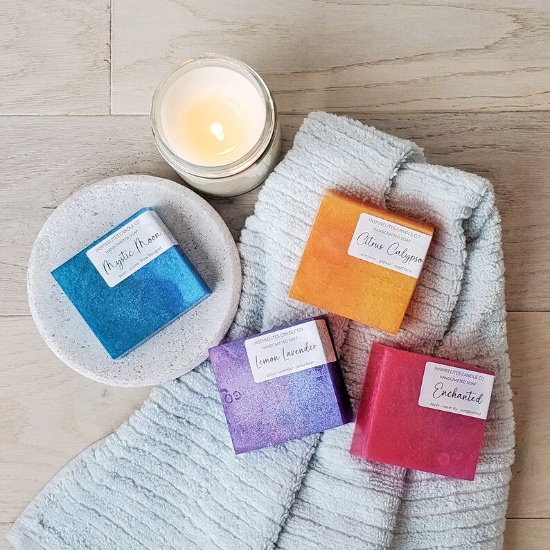 Pode incluir: Quatro barras de sabonete artesanais com diferentes aromas e cores. Os sabonetes est&atilde;o dispostos sobre uma toalha azul com uma vela branca no fundo. Os aromas do sabonete s&atilde;o Mystic Moon, Citrus Calypso, Lemon Lavender e Enchanted.