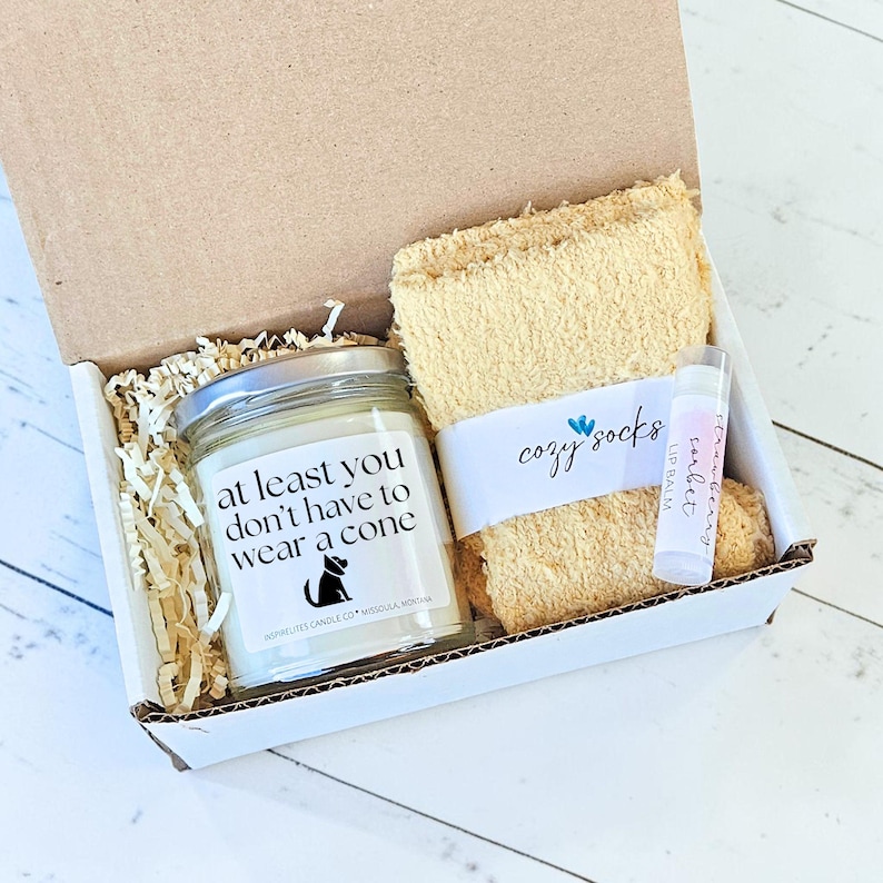 Pu&ograve; includere: Una confezione regalo contenente una candela, calzini e balsamo per le labbra. La candela ha un'etichetta con la scritta "at least you don't have to wear a cone" e una silhouette di gatto. I calzini beige sono etichettati "cozy socks". Il balsamo per le labbra &egrave; etichettato "strawberry".