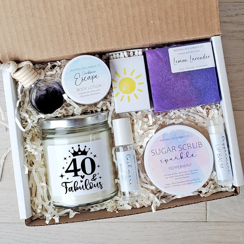 May include: A white gift box filled with self-care items for a 40th birthday. The box includes a candle with the text "40 & Fabulous", a sugar scrub, a bar of soap, a body lotion, a body oil, and a lip balm.