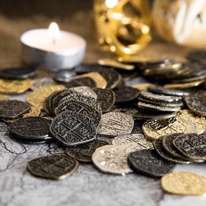 May include: A collection of antique-style coins in various metallic finishes, including gold, silver, and bronze. The coins feature intricate embossed designs and are scattered across a surface, with a lit candle and decorative objects in the background.