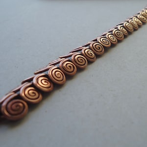 May include: A copper bracelet with a series of spiral-shaped links. The links are arranged in a staggered pattern, with the spirals facing alternating directions. The bracelet is made of a single piece of wire, with the ends joined together.