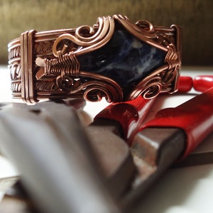 Pode incluir: Um bracelete envolto em fio de cobre com uma pedra preciosa azul e branca engastada no centro. O bracelete está sobre uma superfície branca com um par de alicates vermelhos e prateados.