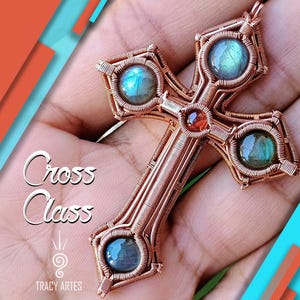 Pode incluir: Um pingente de cruz em fio de cobre com quatro detalhes em pedras preciosas azul-esverdeadas e uma gema vermelha central. A cruz apresenta o texto "Cross Class" e o nome do artista "Tracy Artes".